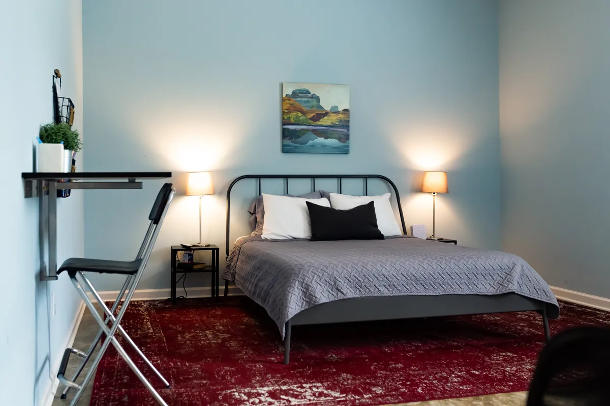A stylishly appointed bedroom with a comfortable bed, creative artwork, soft lighting, and a small desk area by the window. The space offers both a restful sleeping area and a functional workspace.