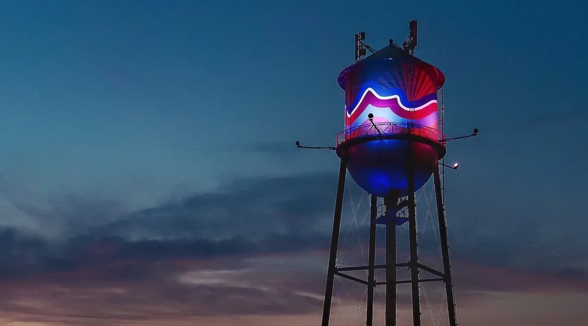 This image features a prominent water tower adorned with colored lights against the evening sky, suggesting a lively community space where people might gather for events or evening walks. The scene conveys a sense of local vibrancy, with the illuminated tower acting as a neighborhood landmark.