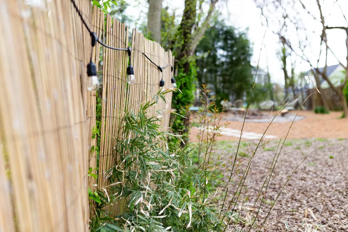 A serene outdoor space defined by a bamboo fence, twinkling hanging lights, and lush greenery. The atmosphere suggests intimacy and warmth, ideal for group or family gatherings in the evening or at dusk.