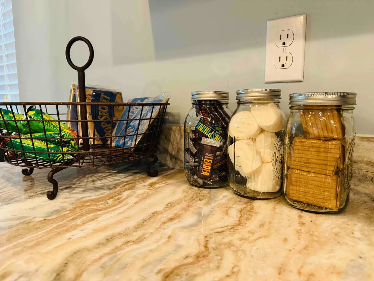 A cozy wire basket brimming with snacks and jars of treats sits in a welcoming interior setting, instantly evoking the fun and communal spirit of gathering for s’mores and shared enjoyment.