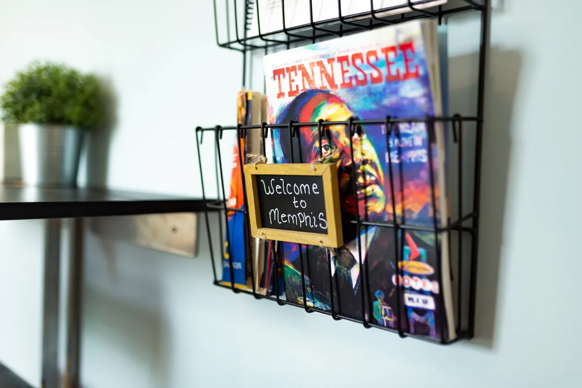 A welcoming interior vignette showcasing a magazine rack filled with colorful local magazines, a prominent welcome sign, and a lush green plant. The scene suggests an inviting atmosphere, perfect for planning outings or engaging with Memphis’s vibrant arts culture.