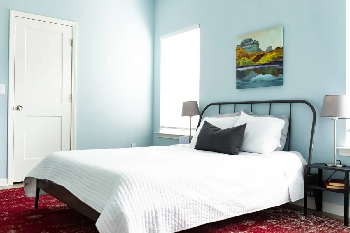 This image highlights a serene bedroom with a bed dressed in a white quilt, colorful painting on the wall, red rug, and ambient table lamps. The room is filled with natural light, creating a comfortable and welcoming atmosphere.
