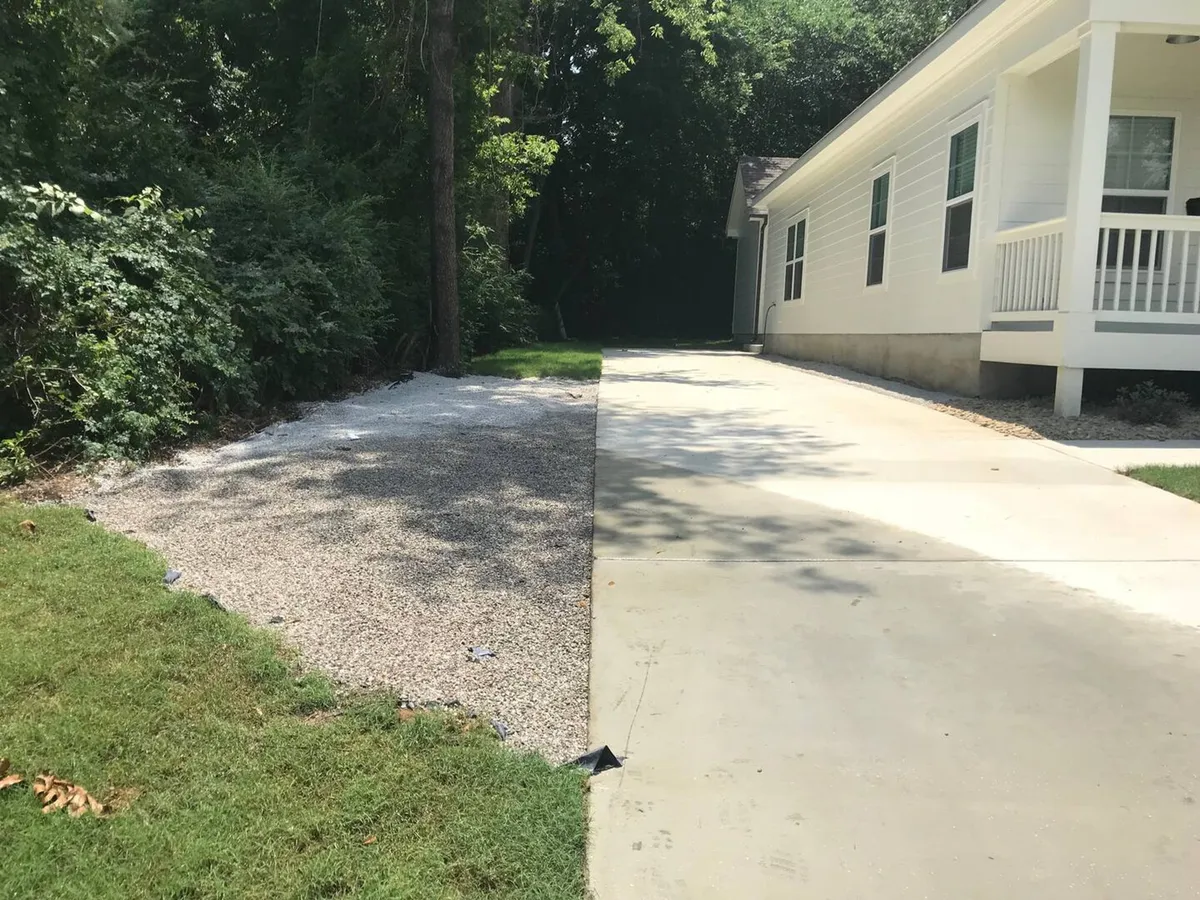A tranquil view of the property’s exterior, highlighting the gravel parking area, concrete driveway, lush grass, and mature trees surrounding the home. The setting is peaceful and expansive, providing a sense of privacy while remaining connected to nature.