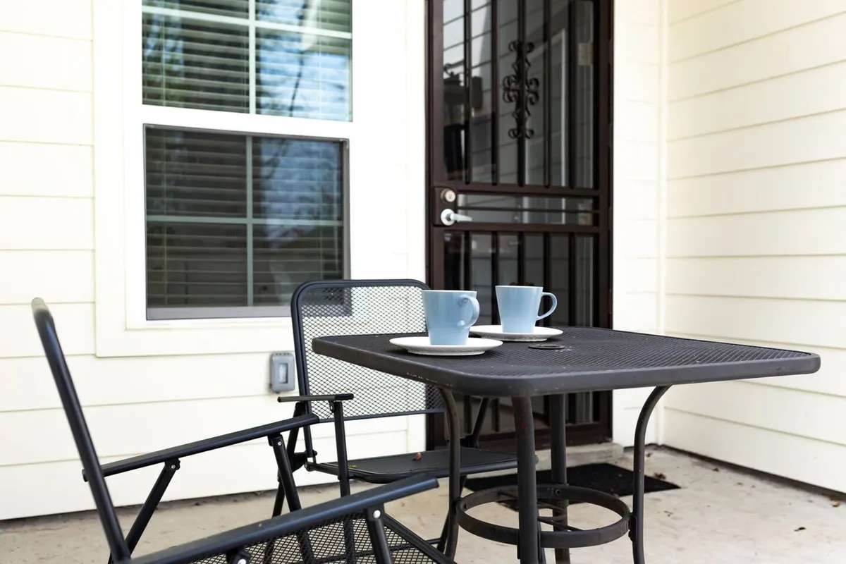 This image captures an inviting exterior space featuring a table and chairs set near the front door, with coffee cups on the table. The scene is bathed in morning sunlight, evoking comfort and a welcoming atmosphere—perfect for both casual work sessions and social gatherings.