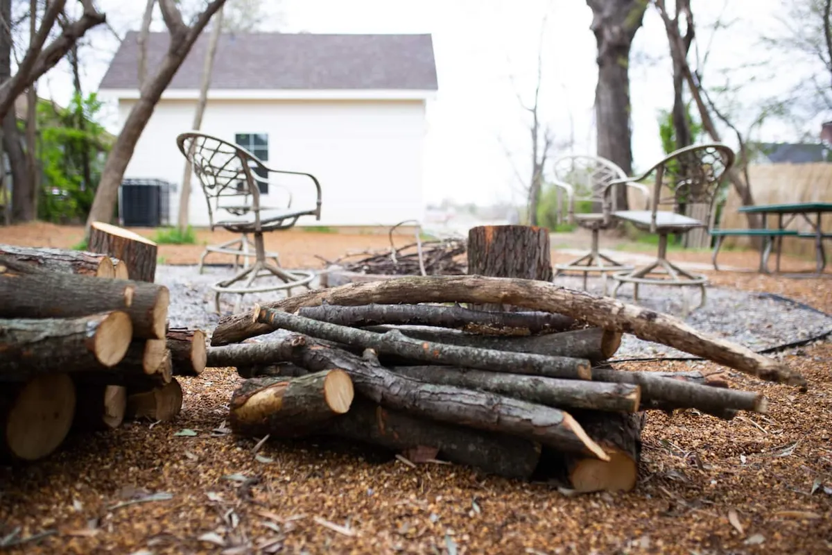 A relaxed outdoor seating area is centered around a fire pit, with a nearby stack of logs ready for the evening's fire. The arrangement invites people to gather and enjoy time together in nature, with comfortable seating and an open sky above.