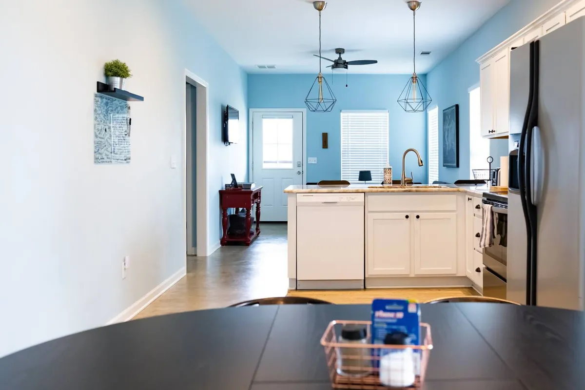A bright, open-plan view encompassing the kitchen island, dining table, and a window that fills the space with natural light. Decorative wall art and an entryway door are also visible, capturing the multifunctional heart of the home ideal for gatherings.